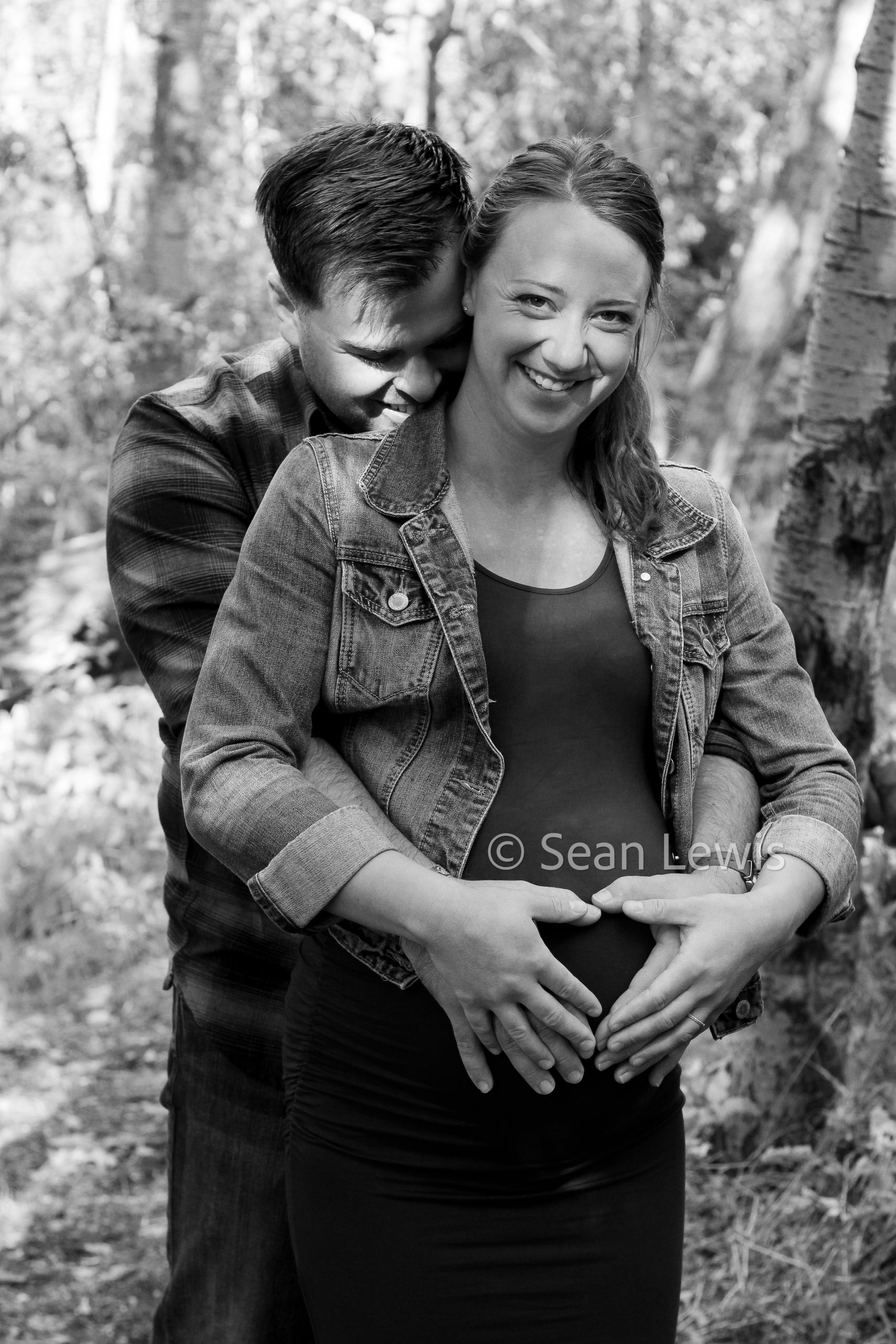 Black and white maternity portrait of a smiling couple with hands on the baby bump in a Wetaskiwin park.